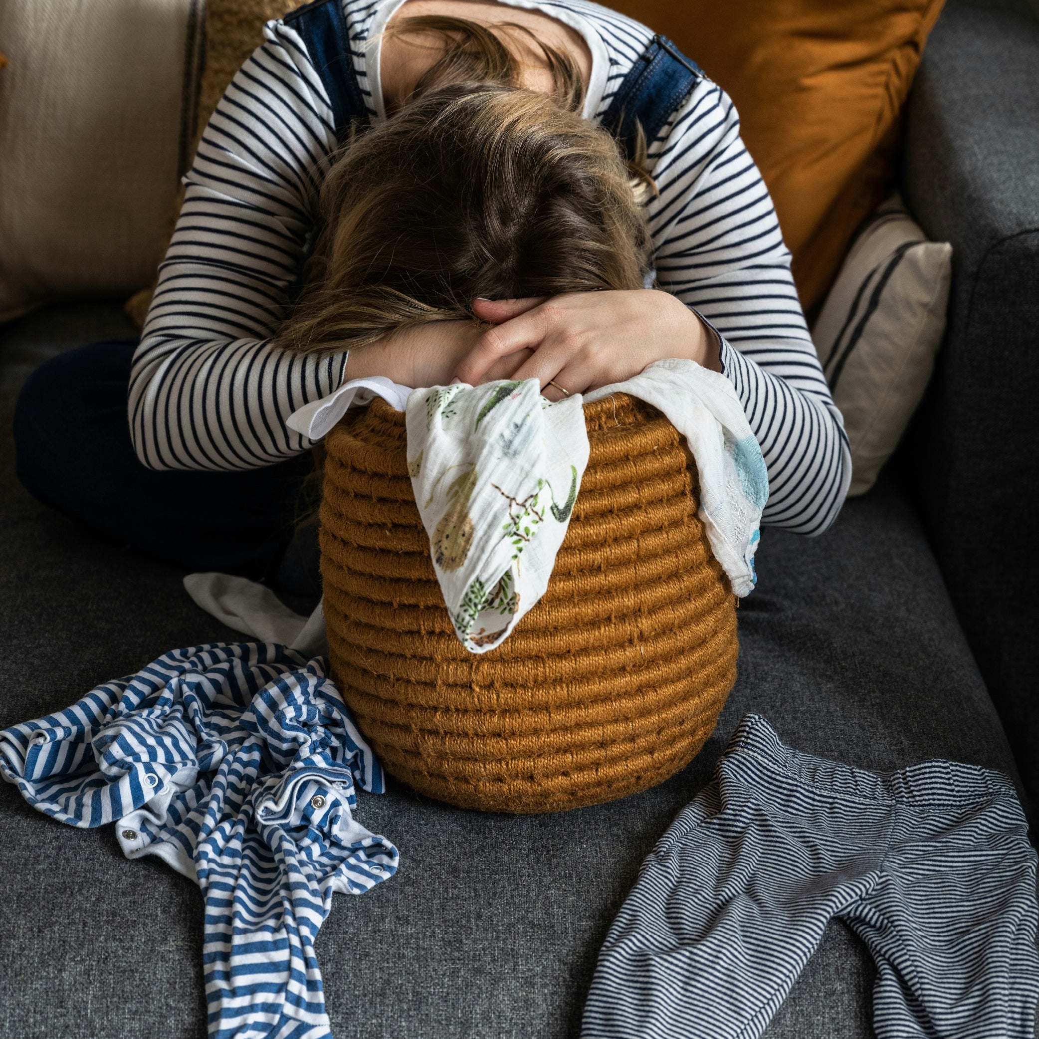 An exhausted mum leans forward with her face hidden in her folded arms, resting on a woven basket of baby laundry on a grey couch