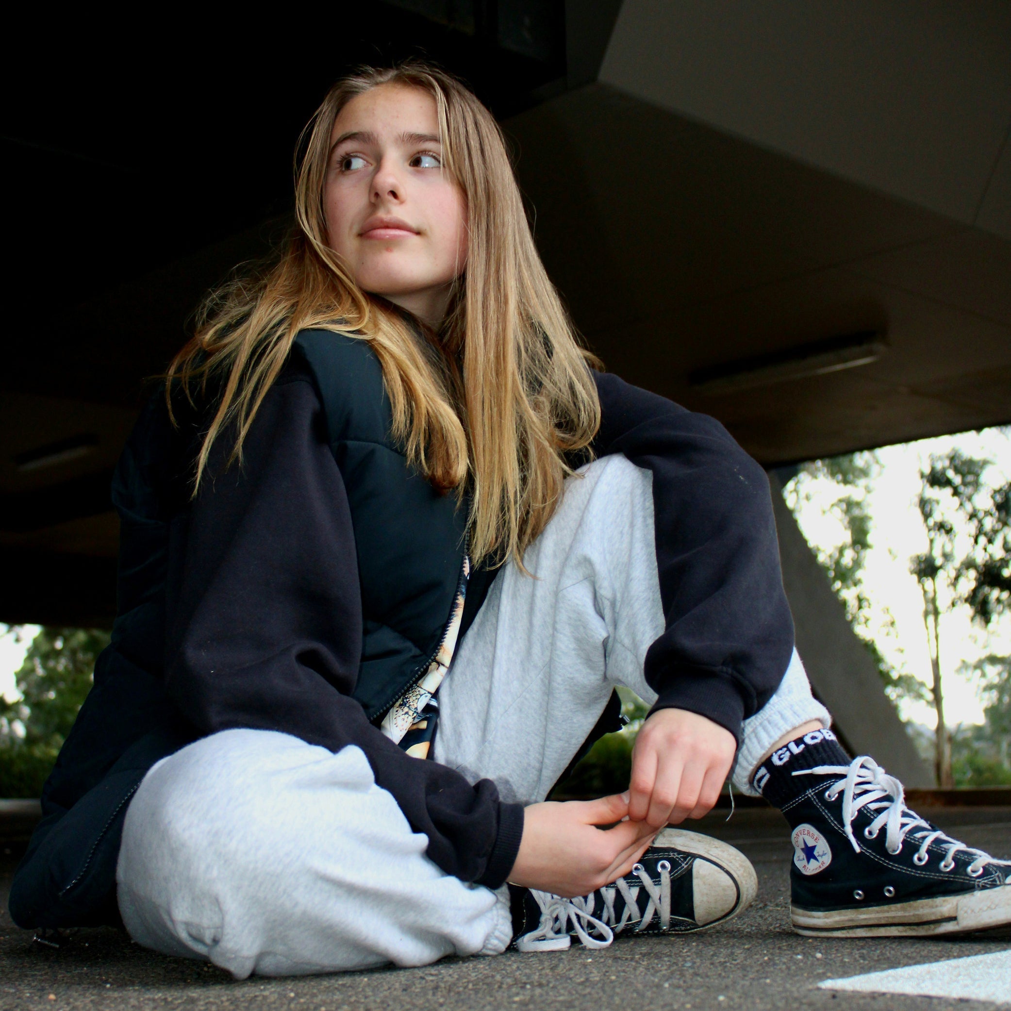A teenage girl sitting cross-legged outdoors in casual winter clothing, looking away from the camera.