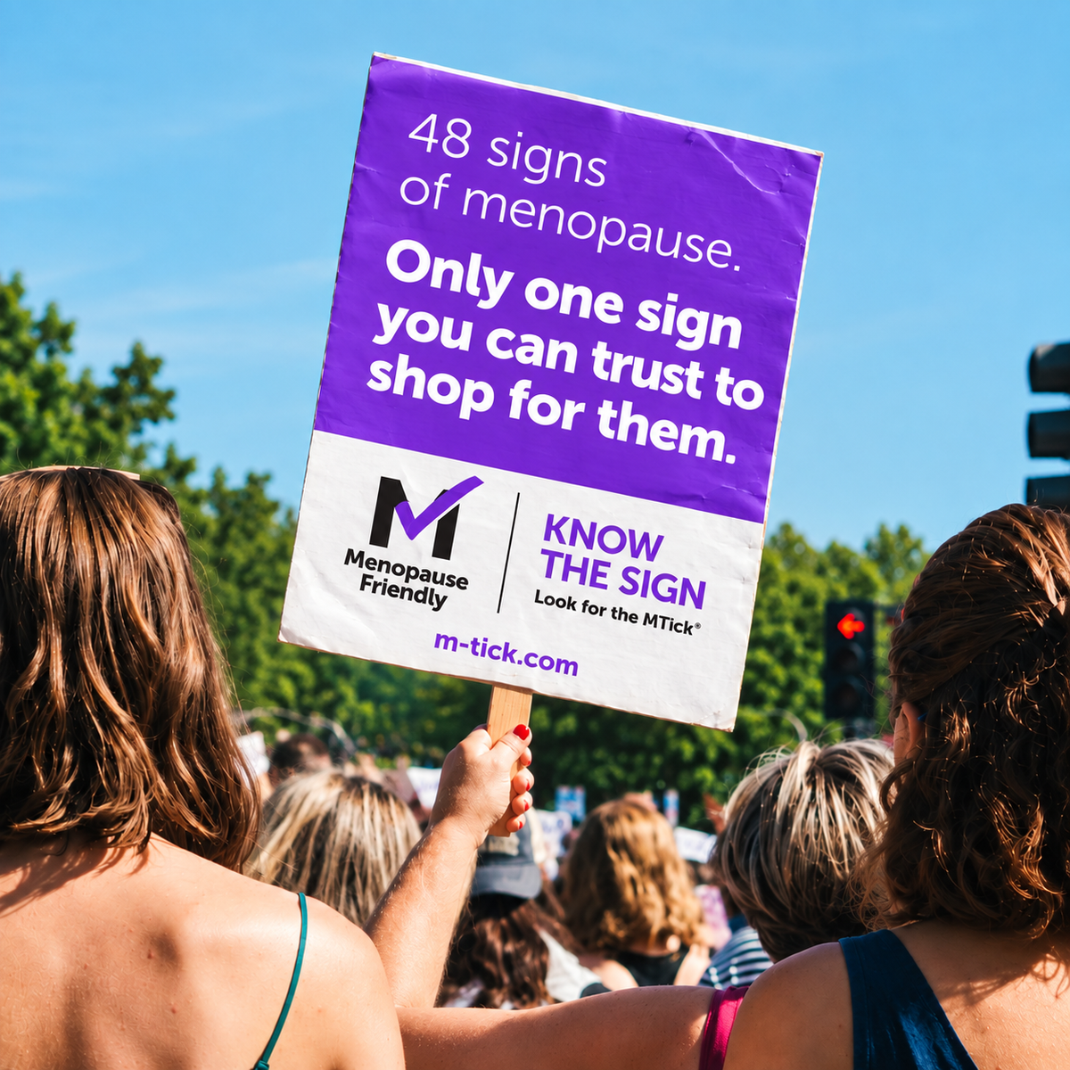 Woman at an outdoor rally holding a purple Menopause Friendly MTick sign reading, “48 signs of menopause. Only one sign you can trust to shop for them.”